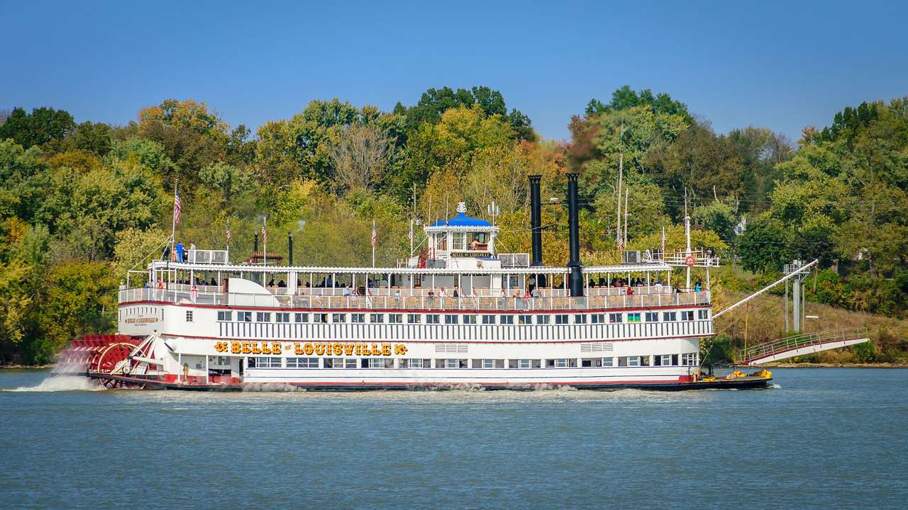 A red and white steamboat on the water next to green trees