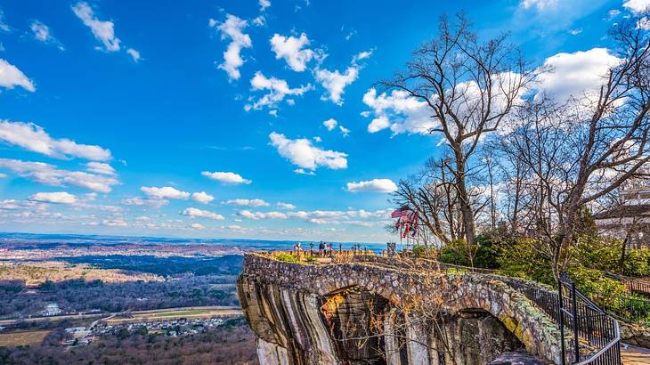 A rocky cliff lookout point with bare trees and greenery below it under a blue sky