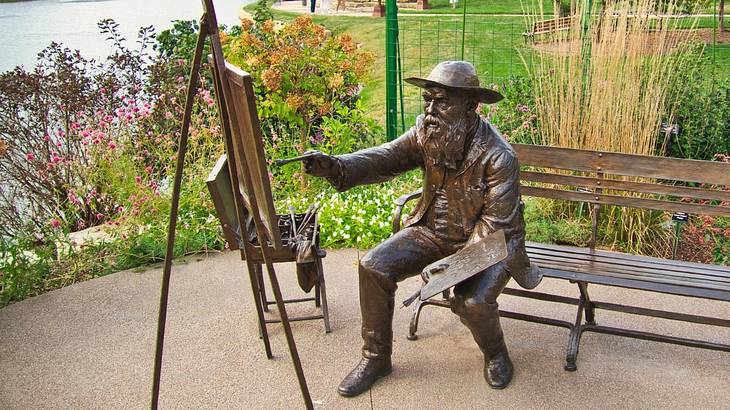 A bronze sculpture of a man on a bench painting on a canvas against greenery
