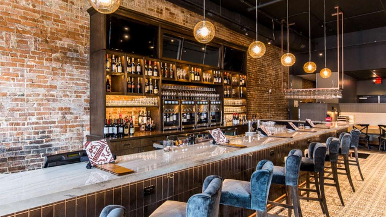 A wine bar with bar stools along the bar and bottles and a brick wall behind it