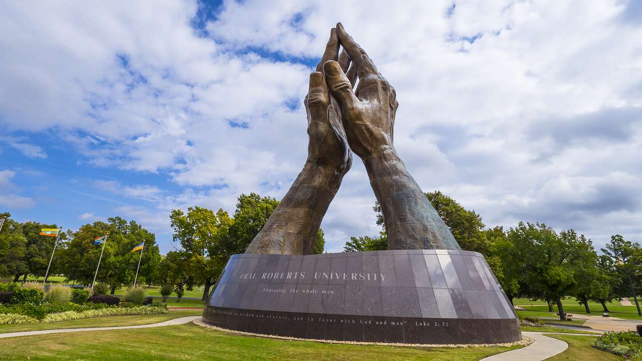 A massive bronze sculpture of praying hands next to the grass and trees