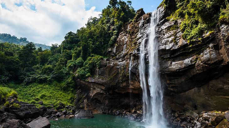 A tall and thin waterfall falling off rock into water below with jungle around