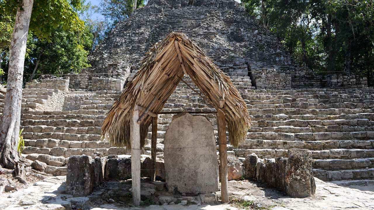 A straw hut housing a big stone slab in front of a large structure with many steps