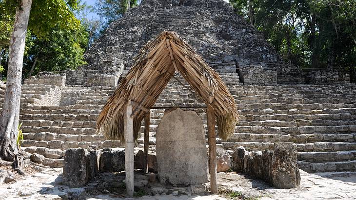 A straw hut housing a big stone slab in front of a large structure with many steps