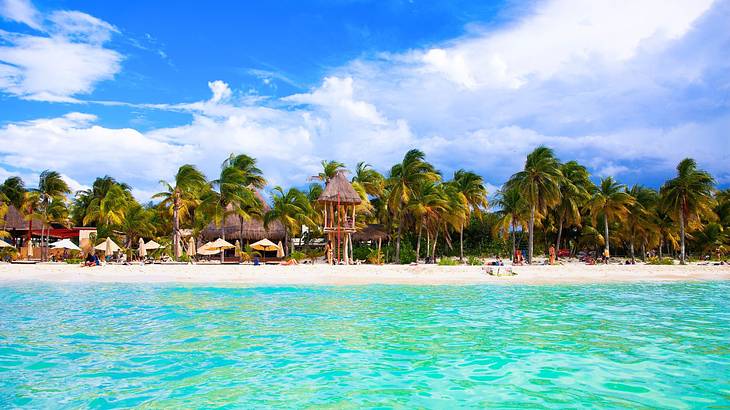 Bright blue ocean water next to a white sand beach with huts and palm trees