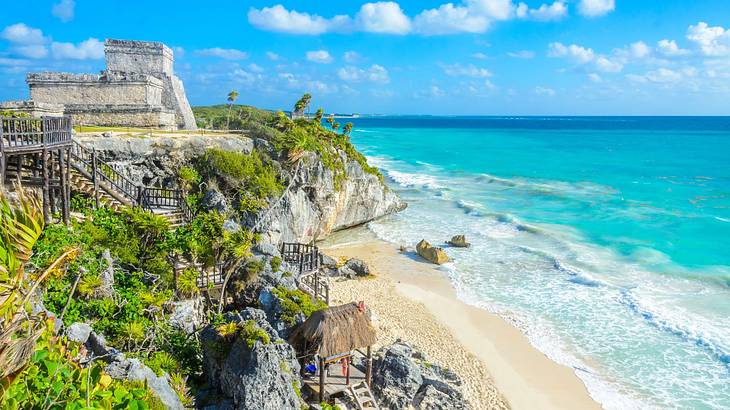 Ancient ruins on a greenery-covered hill next to a sandy beach and turquoise ocean