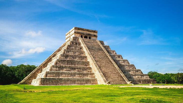 An ancient stone pyramid on the grass next to trees under a blue sky with clouds