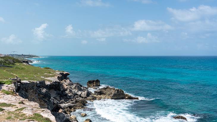 Foamy waves break on a green rocky coast under a blue sky with a few clouds