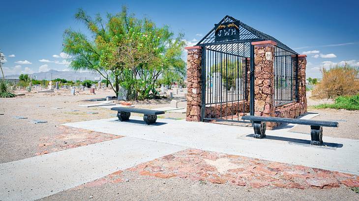 A small jail-like structure near trees, benches, graveyards and a pebbly ground