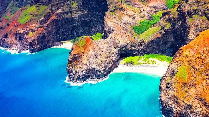 Aerial view of a rocky, reddish coast with lush greenery and calm blue water