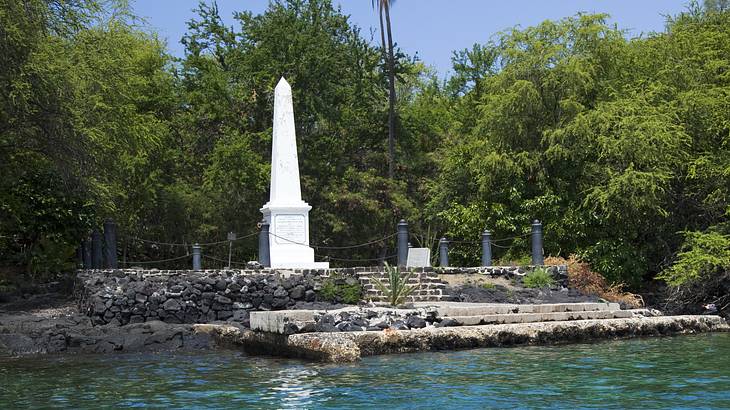 A white obelisk at the side of a river, with green tropical vegetation behind it
