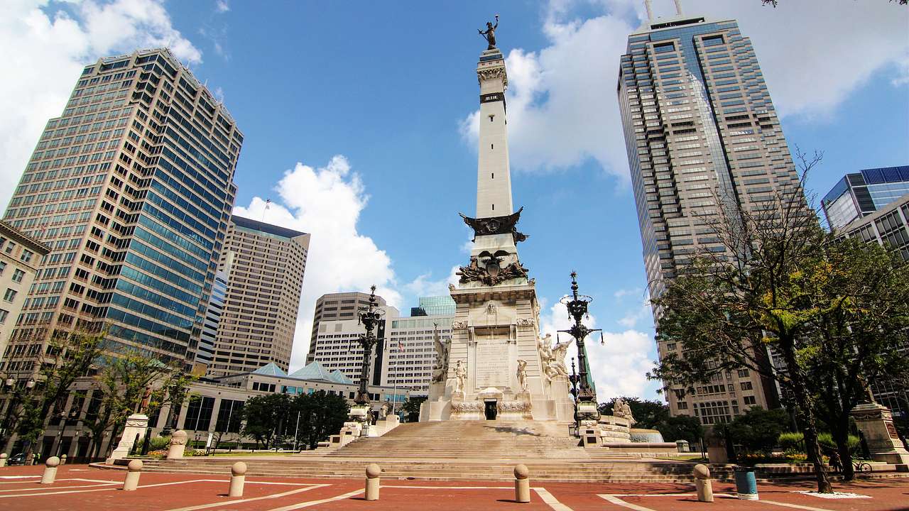 A tall white obelisk adorned with sculptures surrounded by buildings