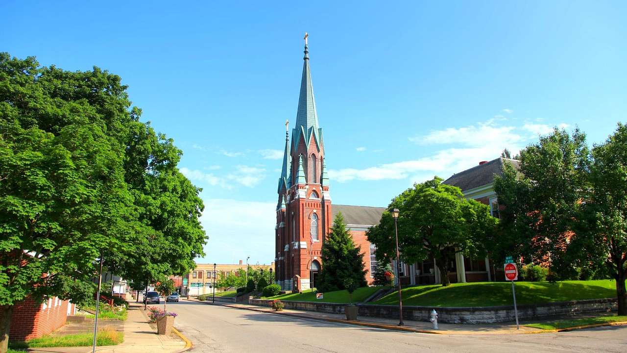 A brick church with a tall spire surrounded by green trees and facing a paved road