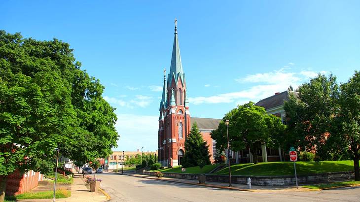 A brick church with a tall spire surrounded by green trees and facing a paved road