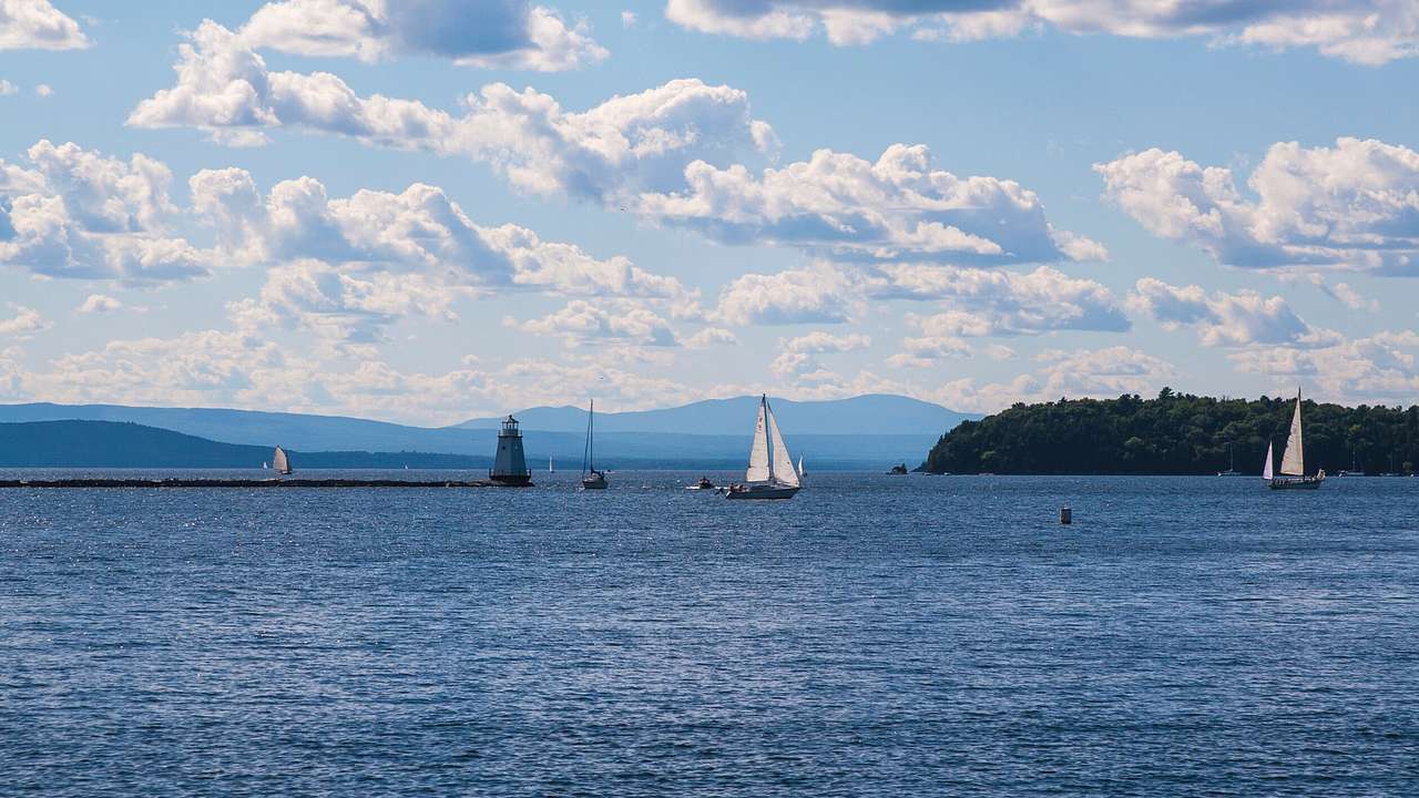 A lake with sailboats and a lighthouse in the distance under a blue sky with clouds