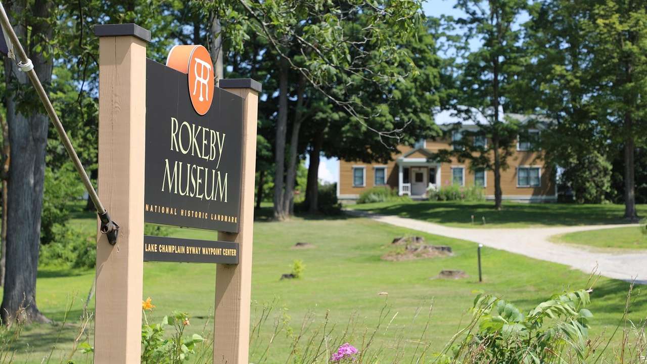 A sign that says "Rokeby Museum" with grass and trees around it and a house behind it