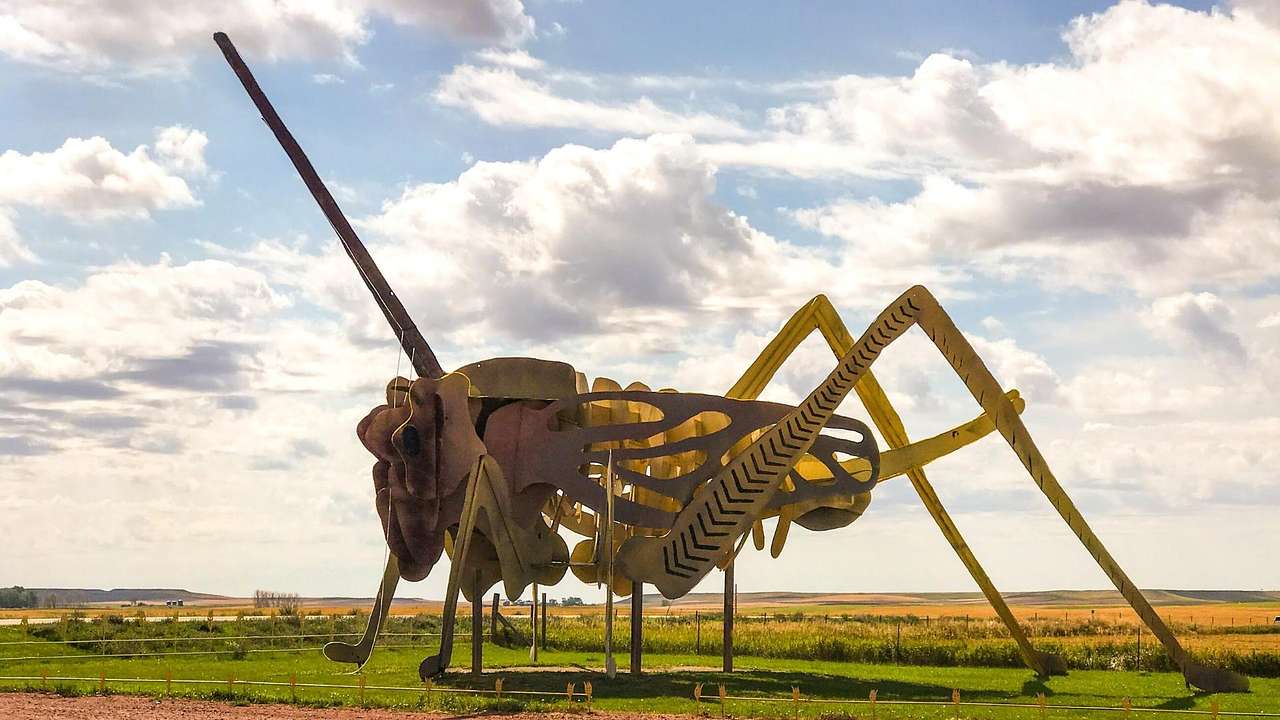 A metal sculpture of a grasshopper on a green field under a partly cloudy sky
