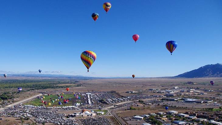 Multi-colored hot air balloons flying over a town with buildings, cars and greenery