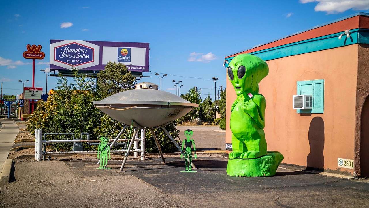 A life-size green alien model and a gray UFO facing a road on a nice day