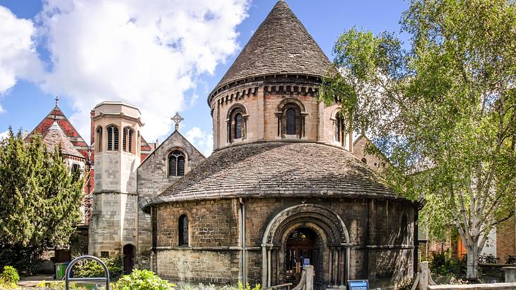 A round medieval church made of stone with a garden, trees, and buildings around