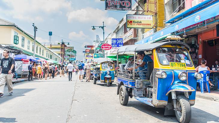 A view of a street lined with shops and people walking, with a blue tuk tuk in front