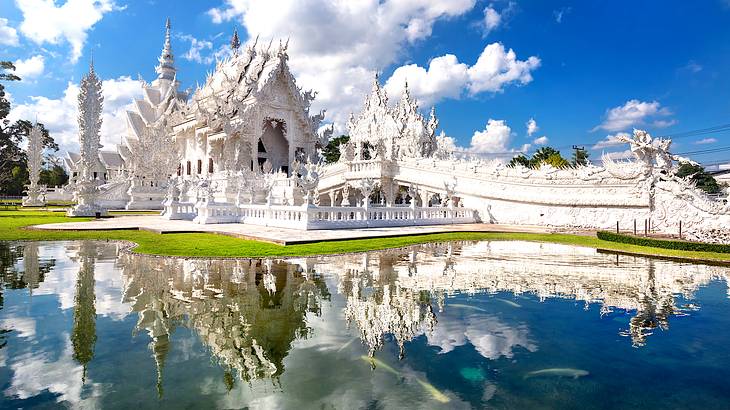 A side view of a detailed white temple with clouds above, reflected in water