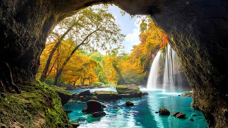 A small waterfall cascading amongst rocks and trees in autumn colours, behind a cave