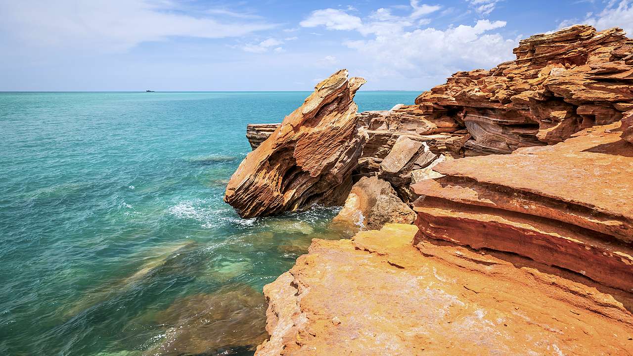 Rocky red cliffs along clear blue water on a partly cloudy day