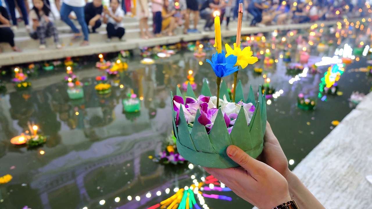 A hand holding a candle with flower petals in front of a canal with floating objects