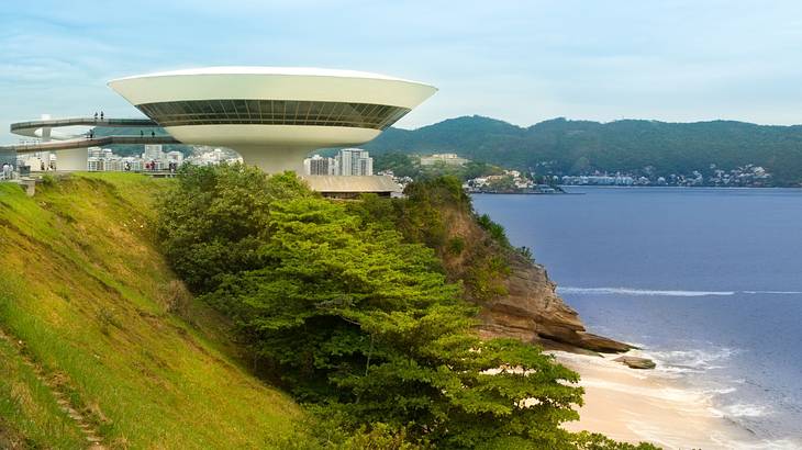 A white spaceship-style building on a green hill overlooking blue water and sand