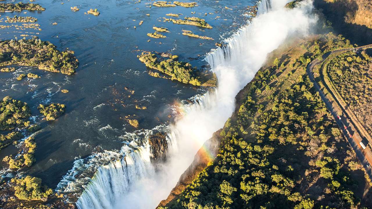 Aerial of a massive waterfall going between lush green cliffs with a rainbow above