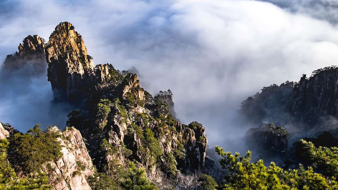 Several rocky mountain summits with trees and bushes, peeking through thick clouds