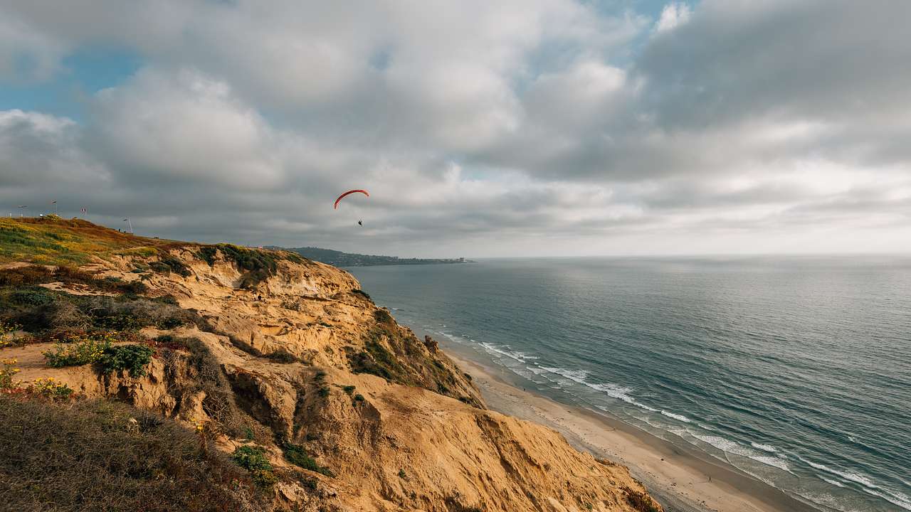 Paraglider flying over a rocky coastline with water below on a cloudy day