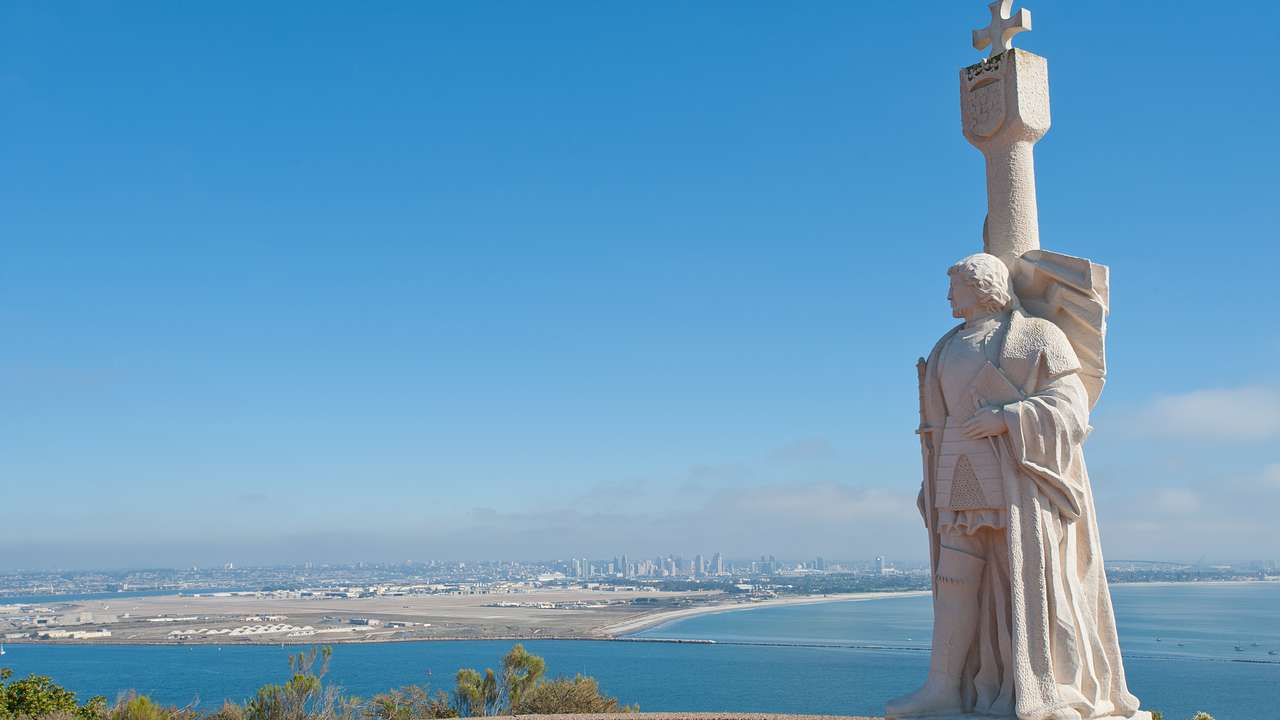 White statue with a cross on top, overlooking the ocean and a city under a blue sky