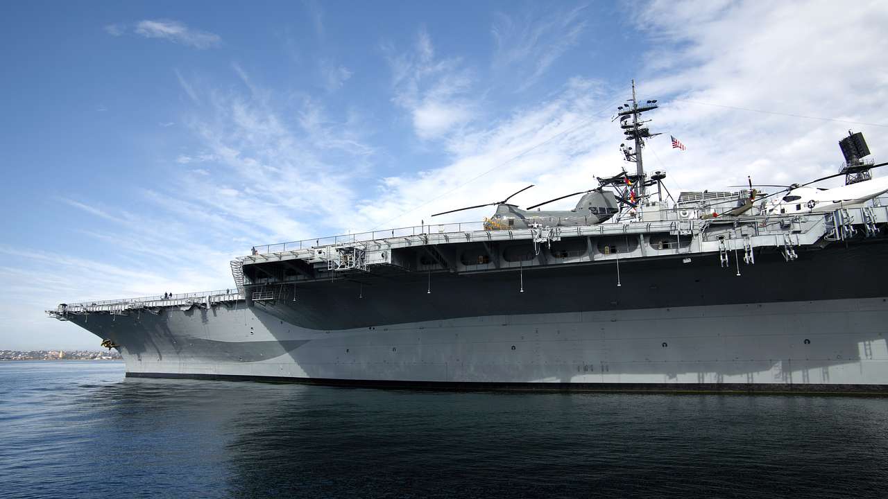 An aircraft carrier with helicopters on top, on a partly cloudy day with water around
