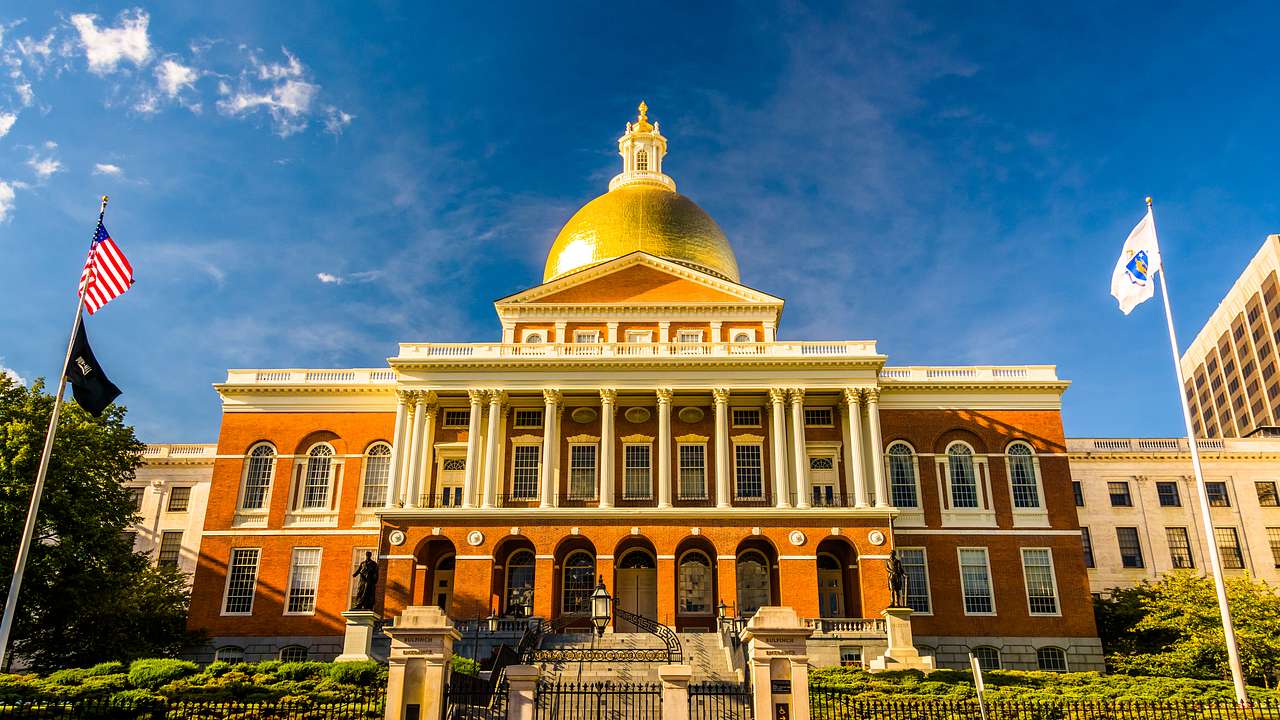 Sun shining on the top of a gold dome building with a fence and green bushes in front