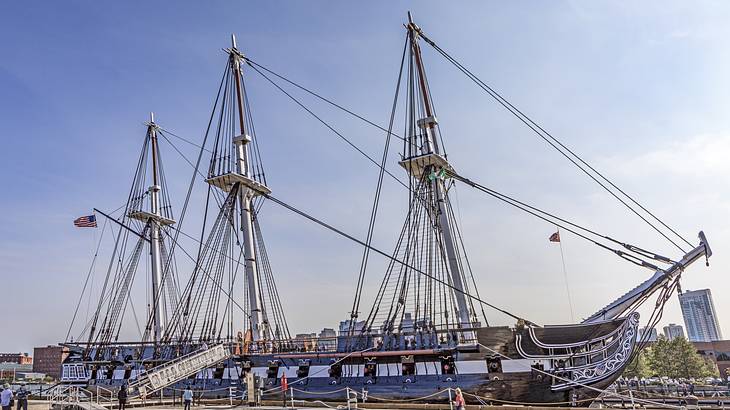 People standing near a three-masted wooden-hulled heavy frigate docked at a pier