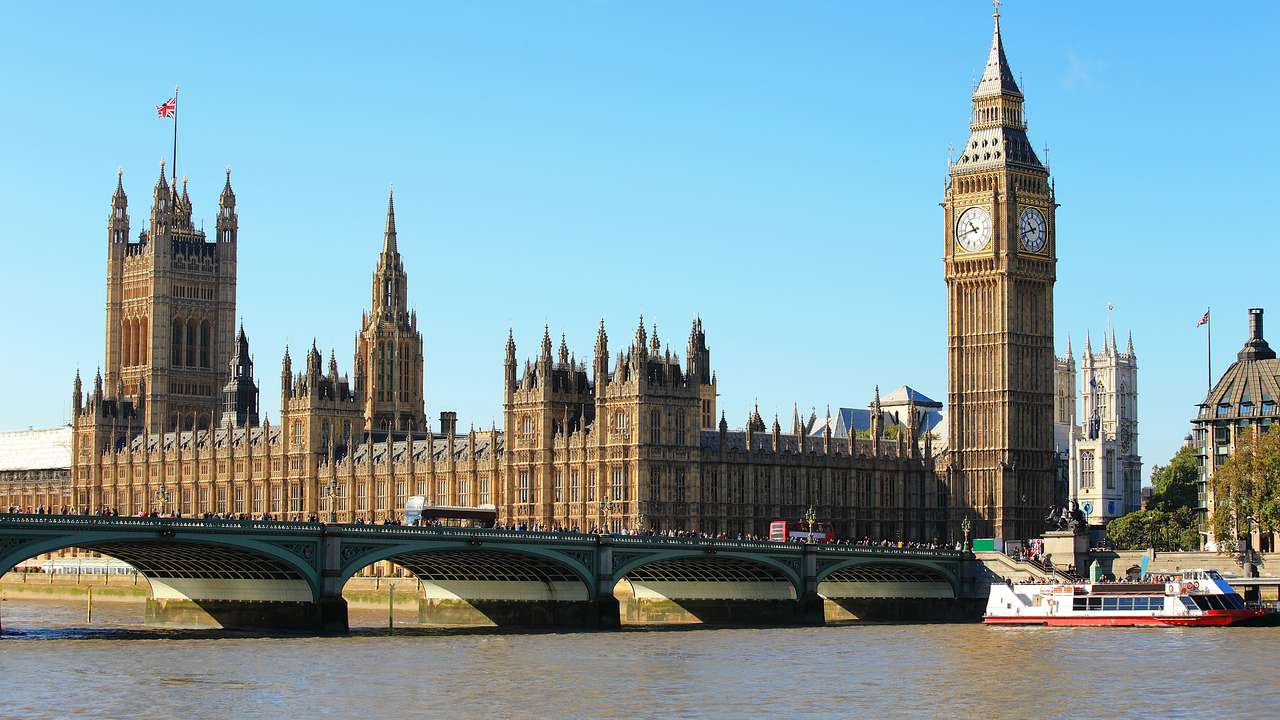 A big palace with a clock tower from across a river, with a bridge and a boat