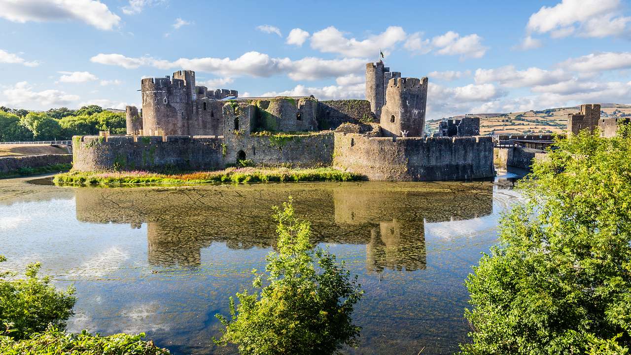 A large stone castle surrounded by clear water and greenery on a sunny day