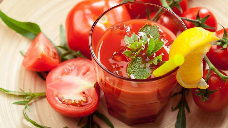 A glass of tomato juice with herbs and a lemon on top, surrounded by tomatoes