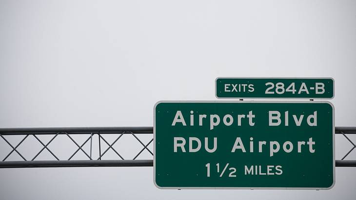 A grey sky behind a green airport highway sign from below