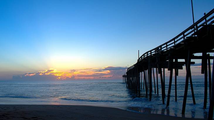 Sunrise over a long fishing pier over water on the right from the shore