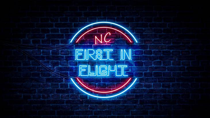 A blue and red LED sign that says "NC FIRST IN FLIGHT" on a dark brick wall