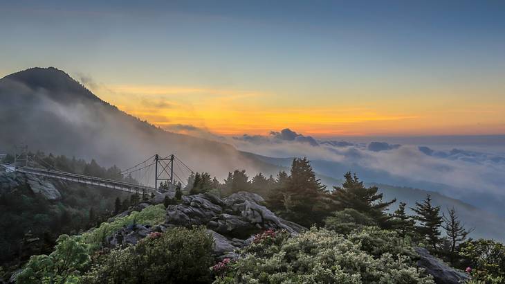 A view of a mountain peak from another with a bridge and clouds at sunrise