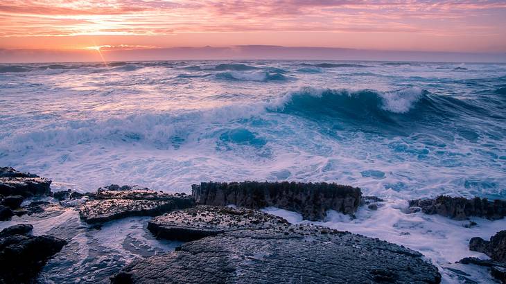 Sunset over the horizon with waves breaking over a rocky coastline
