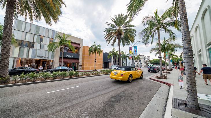 A yellow car driving on a busy street with palm trees on the side and people walking