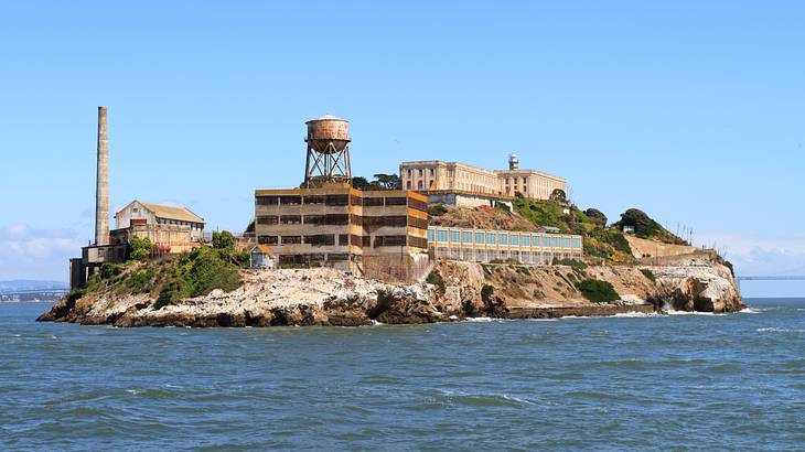 A small hilly island with structures, surrounded by ocean and mountains in the back A small hilly island with structures, surrounded by ocean and mountains in the back
