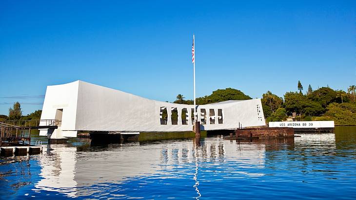 A white memorial, flag, and trees reflected in water against a clear blue sky