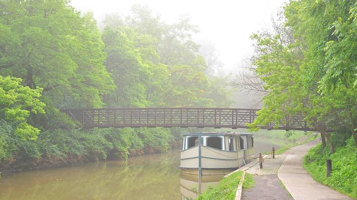 White boat on a canal under a bridge, surrounded by green trees on a foggy day