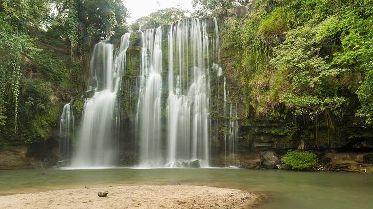 A small sandy beach near the foot of waterfalls, surrounded by greenery